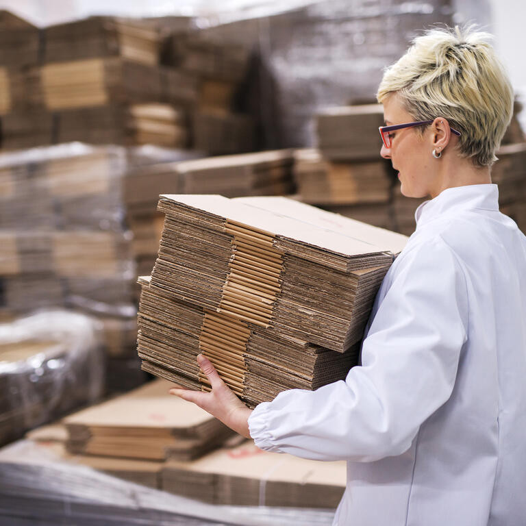 Woman carrying unmade cardboard boxes. Side view, warehouse interior.