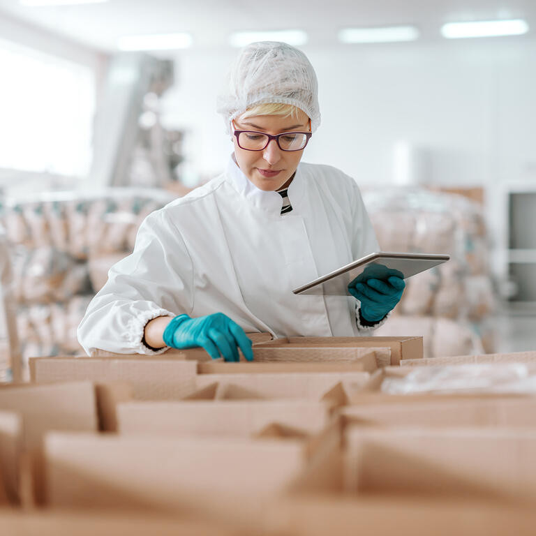 Young Caucasian employee in sterile uniform holding tablet and counting products in boxes. Food factory interior.