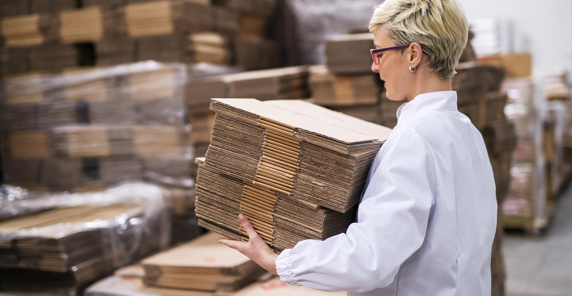 Woman carrying unmade cardboard boxes. Side view, warehouse interior.